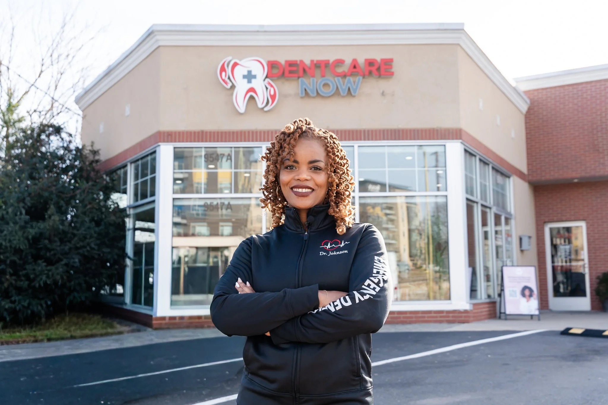 Woman standing with arms crossed in front of Dentcare Now storefront with logo visible